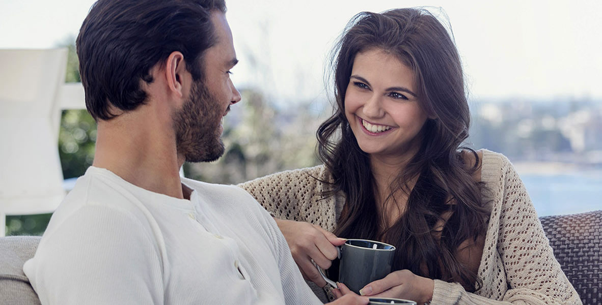 Couple drinking coffee on he sofa. They are sitting on a sofa. They are attractive, smiling and happy. There is a water view from the luxury house.